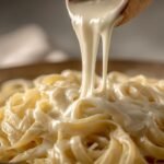Close-up of a wooden spoon pouring rich, creamy alfredo sauce recipe over fettuccine pasta.