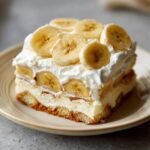 A slice of banana pudding recipe with layers of cookies, pudding, bananas, and whipped cream, topped with fresh banana slices.
