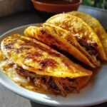 Close-up of three golden-brown birria tacos filled with shredded beef and melted cheese on a white plate.