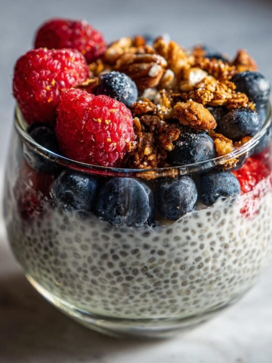 A clear glass filled with creamy chia pudding, topped with blueberries, raspberries, and crunchy granola.