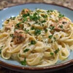 A close-up of a bowl of fettuccine pasta coated in creamy Alfredo sauce with chunks of seasoned chicken and fresh parsley.