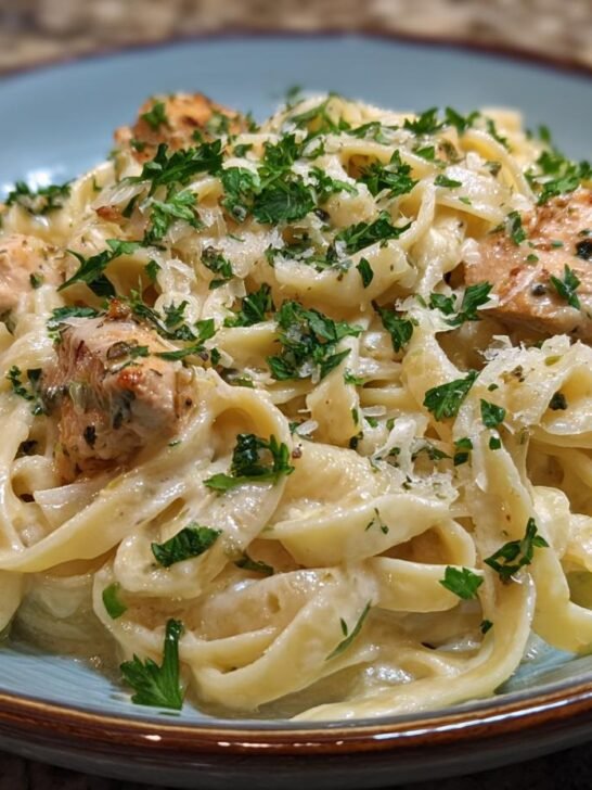 A close-up of a bowl of fettuccine pasta coated in creamy Alfredo sauce with chunks of seasoned chicken and fresh parsley.