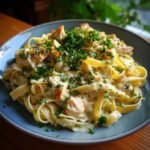 A close-up of a serving of fettuccine pasta with creamy sauce and chunks of chicken, garnished with fresh parsley. This is a chicken alfredo recipe.