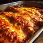 Close-up of a baking dish filled with golden-brown chicken enchiladas, topped with bubbling melted cheese and red enchilada sauce.