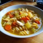 A close-up of a bowl of chicken noodle soup, featuring pasta, shredded chicken, carrots, and celery in a savory broth.
