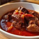 A close-up shot of a rich and savory goulash recipe in a white bowl, featuring tender chunks of beef in a deep red sauce with onions and tomatoes.