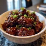 A bowl of Irresistible Asian Crockpot Meatballs coated in a glossy sauce, sprinkled with sesame seeds and chopped green onions.