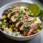 Close-up of a bowl filled with rice, black beans, corn, and sliced honey chipotle chicken, garnished with cilantro and lime.