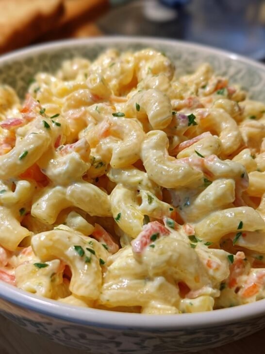 A close-up of a bowl filled with a creamy macaroni salad recipe, garnished with chopped parsley and bits of red pepper.