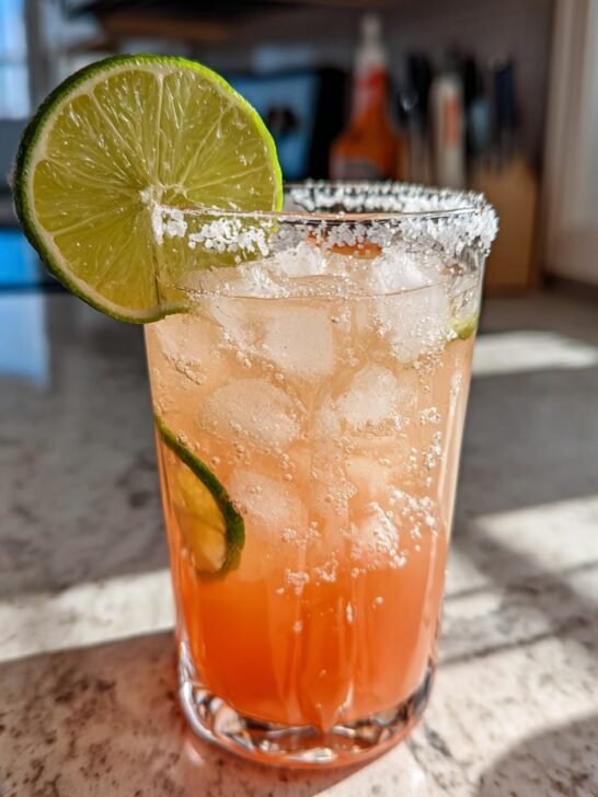 A close-up of a refreshing paloma recipe cocktail, garnished with a lime wedge and salt rim, filled with ice and bubbles.