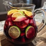 A clear glass pitcher filled with a vibrant red sangria recipe, packed with ice, orange slices, lime slices, and apple chunks.