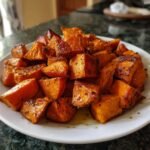 A close-up shot of roasted sweet potato cubes piled high on a white plate, glistening with juices.