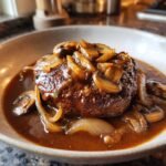 Close-up of a juicy Salisbury steak recipe topped with sautéed mushrooms and onions in a rich brown gravy.