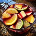 Close-up of a glass pitcher filled with a vibrant red sangria recipe, featuring slices of oranges, limes, and chunks of fruit.