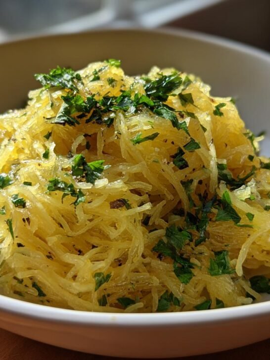 A close-up of a white bowl filled with cooked spaghetti squash, seasoned and topped with fresh parsley.