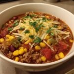 A close-up of a white bowl filled with a hearty taco soup recipe, topped with shredded cheese and cilantro.