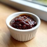 A close-up of thick, dark, glossy Homemade BBQ Sauce served in a small white ramekin.