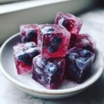 A small stack of vibrant, reddish-purple Blueberry Lemonade Ice Cubes, each containing whole blueberries, sitting in a white ceramic bowl.