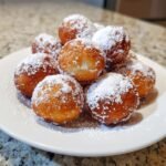 A pile of freshly fried Bomboloni, dusted generously with powdered sugar, on a white plate.