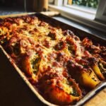 A close-up, overhead view of baked Broccolini And Jerk Chicken Stuffed Shells in a baking dish, topped with melted cheese.
