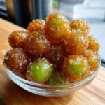 A close-up of a glass bowl filled with glistening Candied Frozen Sour Grapes, coated in a sweet syrup and sugar crystals.