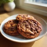 Three golden brown, chewy brown butter pecan cookies stacked slightly on a light-colored plate near a window.