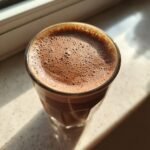 A close-up of a glass filled with frothy Chocolate Almond Milk, catching the sunlight.