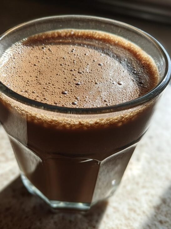 A close-up of a glass filled with frothy Chocolate Almond Milk, with sunlight casting shadows.