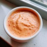 A close-up overhead shot of thick, orange-pink Creamy Thousand Island Dressing speckled with red spices in a small white ramekin.