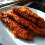 Close-up of several golden, crispy buffalo chicken tenders coated in a shiny, spicy orange sauce on a white plate.