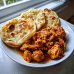 A plate featuring rich, saucy Curry Chicken Roti served with three pieces of golden, flaky flatbread.