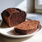 A loaf and a slice of moist Easy Vegan Chocolate Bread displayed on a white plate in natural light.