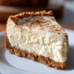 A close-up of a creamy slice of Eggnog Cheesecake Snickerdoodle Crust dusted with cinnamon.