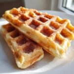 Two golden, fluffy cinnamon vegan Belgian waffles dusted heavily with cinnamon sugar on a white plate.