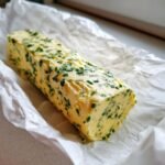 A log of homemade Garlic Herb Butter studded with bright green chives, resting on crumpled white parchment paper.