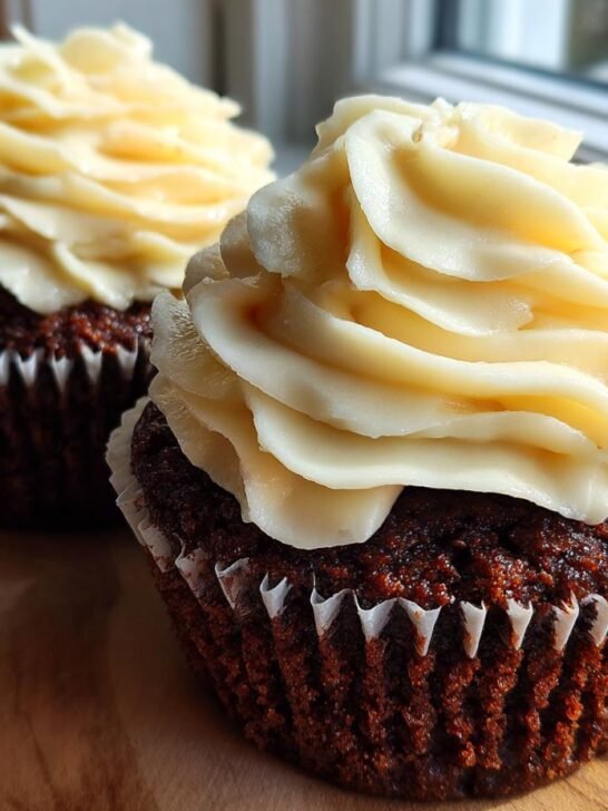 Two rich Gingerbread Cupcakes Eggnog Frosting treats sitting on a wooden surface near a window.