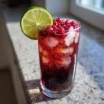 A tall glass filled with a fizzy, deep red Holiday Pomegranate Ginger Mocktail, ice, and topped with pomegranate seeds and a lime wheel.