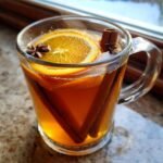 A warm glass mug filled with Homemade Slow Cooker Apple Cider, garnished with orange slices, cinnamon sticks, and star anise.