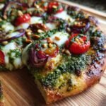 A close-up of a slice of Kale Pesto Pizza topped with cherry tomatoes, red onion, mozzarella, and pine nuts.