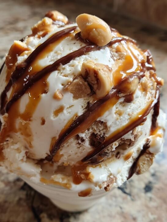 A close-up of a scoop of Loaded No Churn Snickers Ice Cream, drizzled with chocolate and caramel, and topped with peanuts.