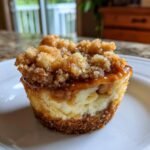 A close-up of a single Mini Salted Caramel Apple Streusel Cheesecake Bite on a white plate, showcasing the creamy cheesecake filling, caramel drizzle, and streusel topping.