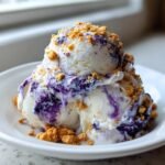 Two scoops of No Churn Blueberry Protein Ice Cream swirled with purple blueberry sauce and topped with gingersnap crumbs.
