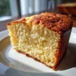 A close-up, angled view of a slice of moist Olive Oil Cake on a white plate, showing its tender crumb and golden-brown crust.