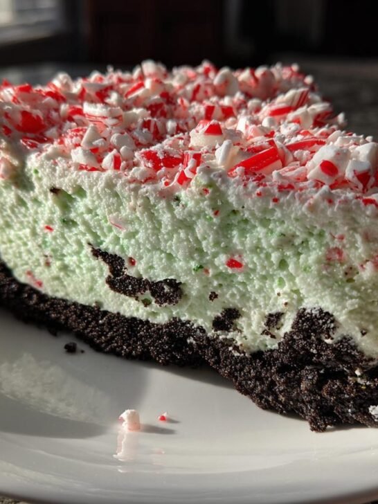 A close-up slice of light green Peppermint Oreo Cheesecake with a dark chocolate crust, topped with crushed candy canes.