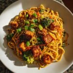 A close-up of Plant Based Sesame Chicken Noodles featuring glazed tofu cubes, broccoli florets, and shredded carrots, topped with sesame seeds.