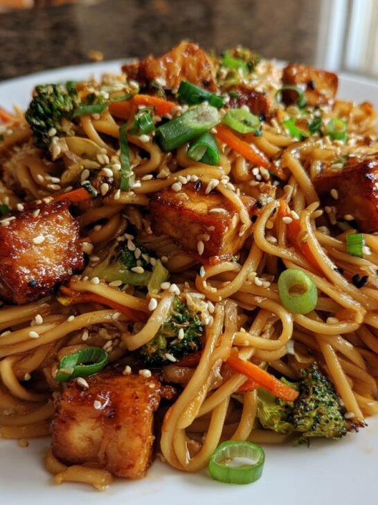 A close-up of delicious Plant Based Sesame Chicken Noodles topped with crispy tofu, broccoli, and sesame seeds.