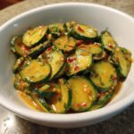 Close-up of sliced cucumbers coated in a vibrant, spicy peanut sauce with chili flakes and green onions.