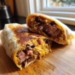 A grilled Ribeye Steak Burrito cut in half, revealing steak, rice, and black beans inside, resting on a wooden board.