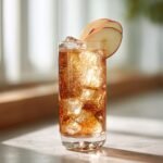 A tall glass filled with ice and a sparkling amber Roasted Apple Pear Mocktail, garnished with thin apple slices.