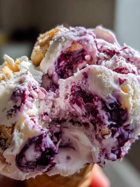 A close-up of a scoop of Southern Blackberry Cobbler Ice Cream in a cone, showing swirls of blackberry and chunks of cobbler.
